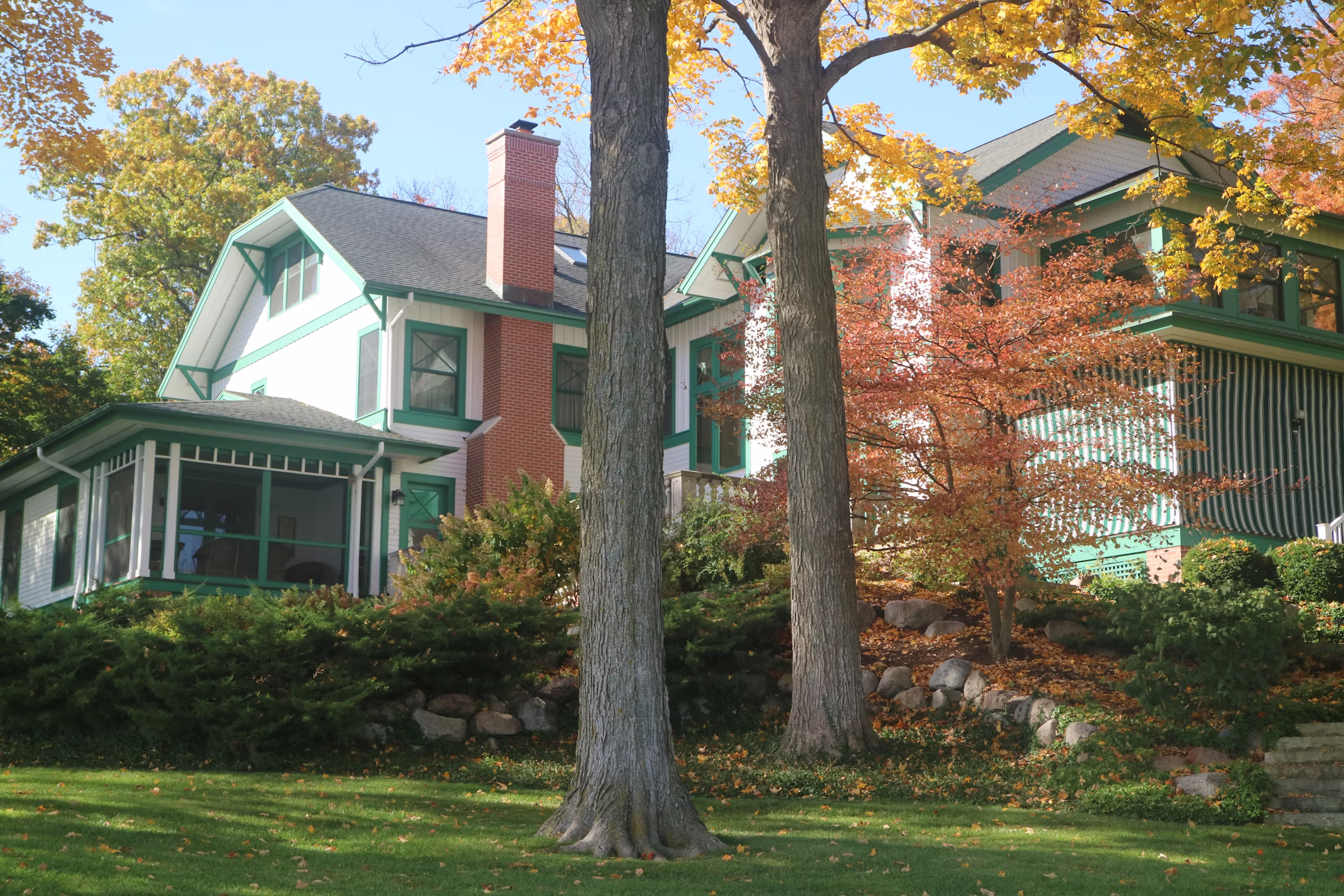 A Folk Victorian-style home with white siding, green trim, brick chimney, and mature trees in autumn color