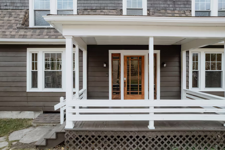 A covered front porch with white railings and columns, a warm wood-toned front door, and dark charcoal siding — the entry detail of a home under inspection