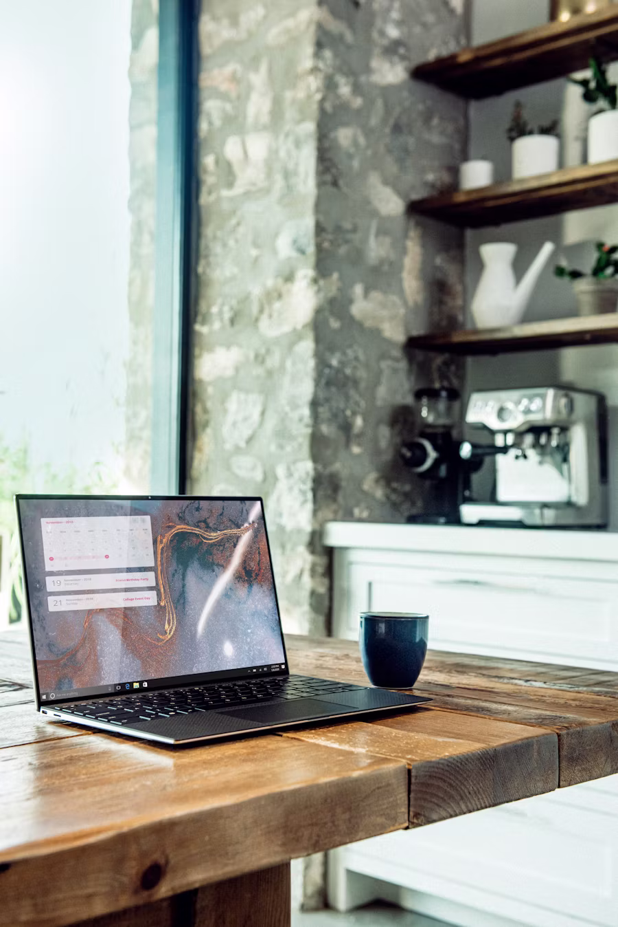 A laptop open on a rustic wooden table next to a coffee mug, with an espresso machine in the background — the editorial-workspace energy of running the math yourself