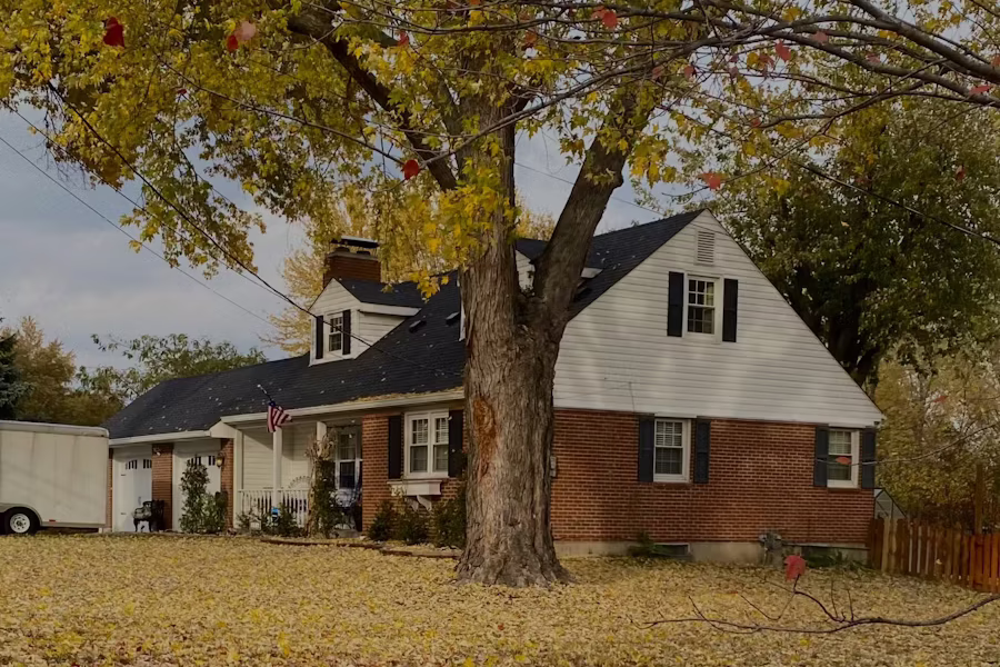A brick and white-trim home framed by an autumn tree with fallen leaves on the lawn — a residential streetscape in Westland, Michigan