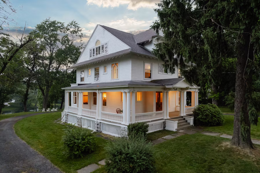 Folk Victorian home at twilight with wraparound porch and lit interior — Wixom's older housing stock